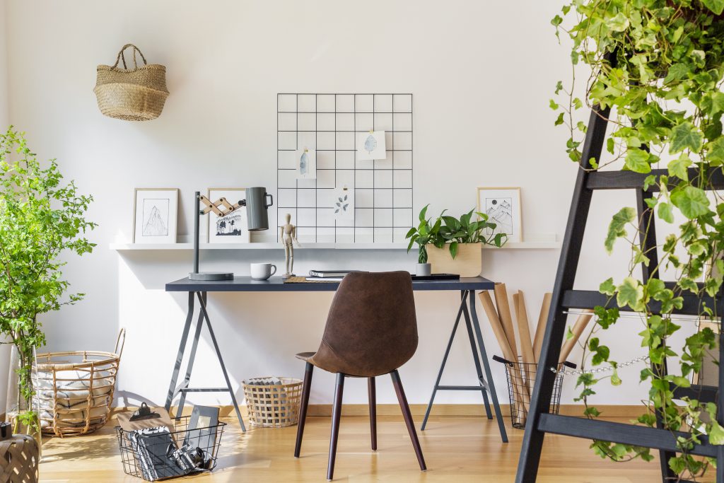 Brown chair at desk in white boho home office interior with plants and posters.