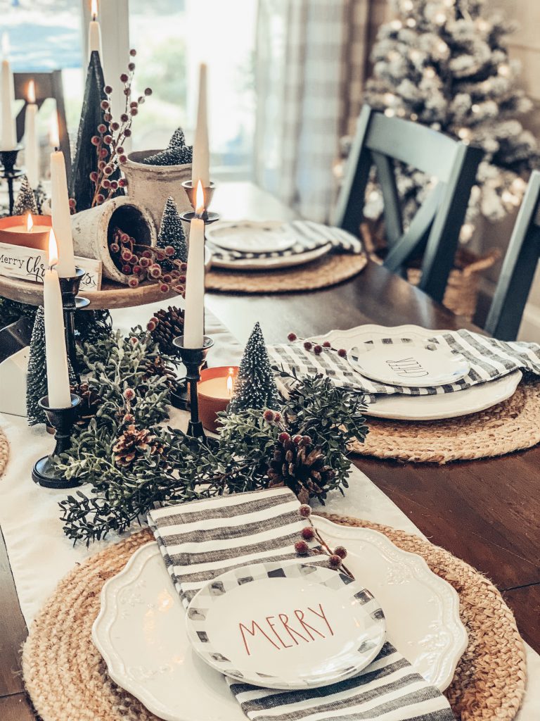 A holiday tablescape with Rae Dunn plates, candles, and greenery with pinecones and red berries
